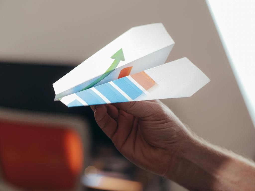 A hand holding a colorful paper airplane with a green upward arrow design.
